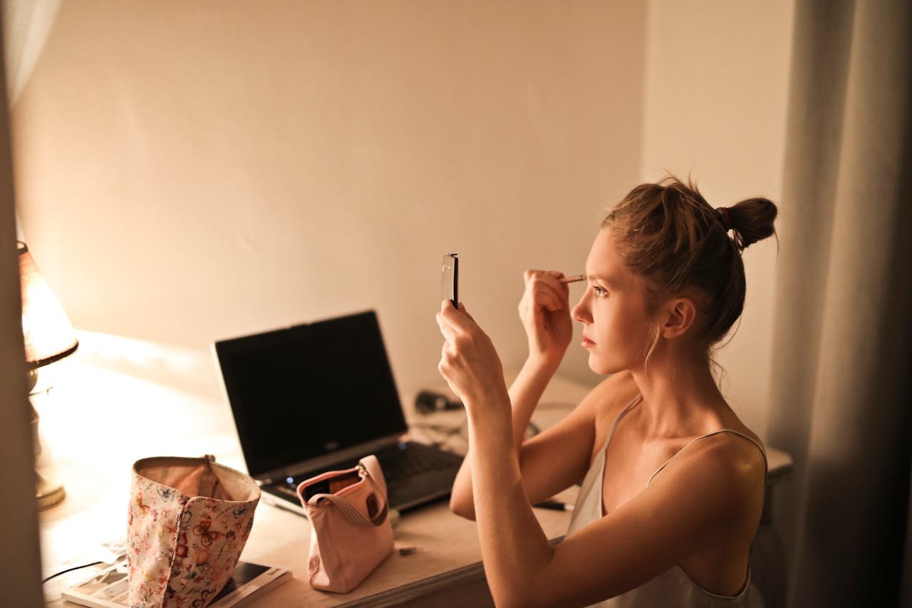 creative-02 A young woman applies makeup at her vanity table, illuminated by soft light, in a cozy home setting.